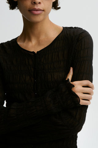 By Malene Birger Ebba Cardigan in black, featuring a unique smocked and puckered elasticated finish with delicate ruffle trim. This sheer, lightweight long-sleeve top offers a slim fit and feminine texture for transitional layering.